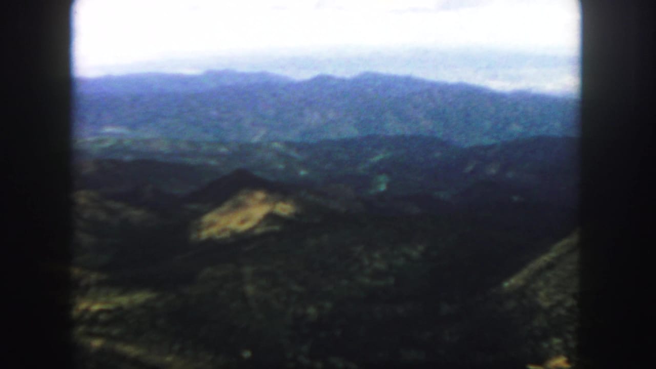 A broad mountain panorama with blue ridges, dark foreground slopes, and bright sky.