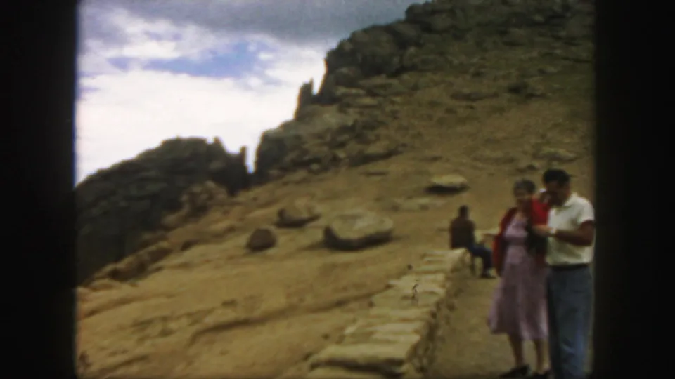 People on a Colorado mountain summit path, including a woman in a pink dress and a man in a white shirt, with rocks behind them.