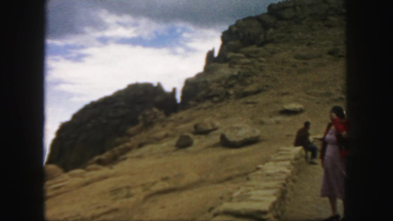 1958 Colorado home-movie frame showing a rocky dirt hillside, clouds, and small visitors near a stone retaining wall.
