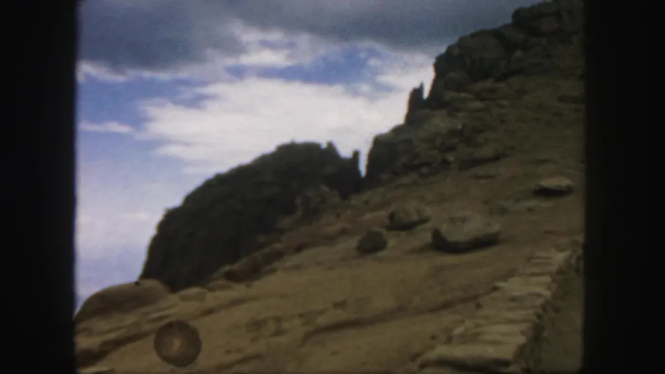 1958 Colorado frame of a sloped rock formation beneath clouds, with the road edge and a small round object visible below.