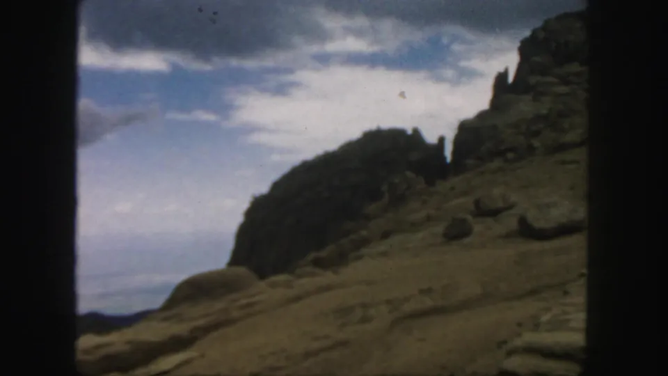 1958 Colorado landscape frame with boulders, a slanted dirt slope, and clouds visible through faded 8mm color.