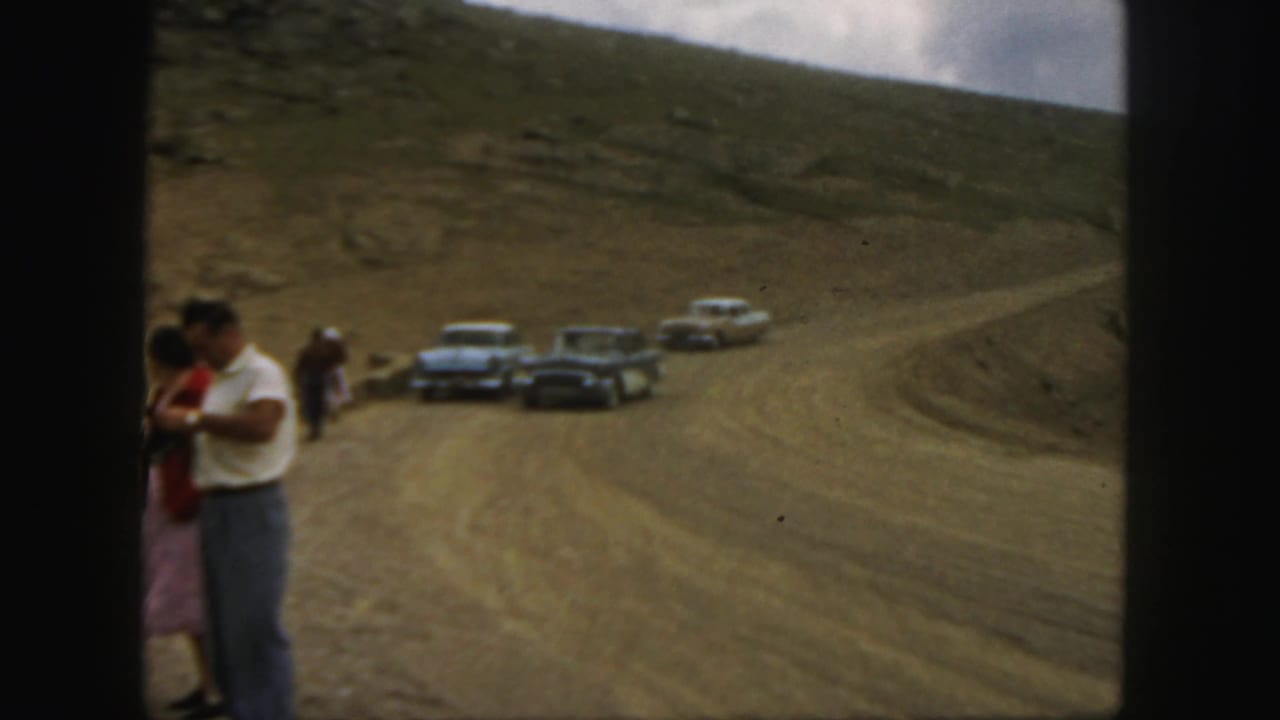 1958 Colorado home-movie frame with a couple at roadside and several classic cars on a winding dirt mountain road.