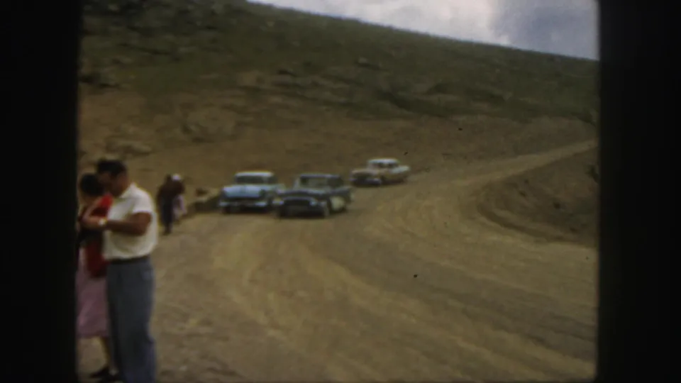 1958 Colorado home-movie frame with a couple at roadside and several classic cars on a winding dirt mountain road.