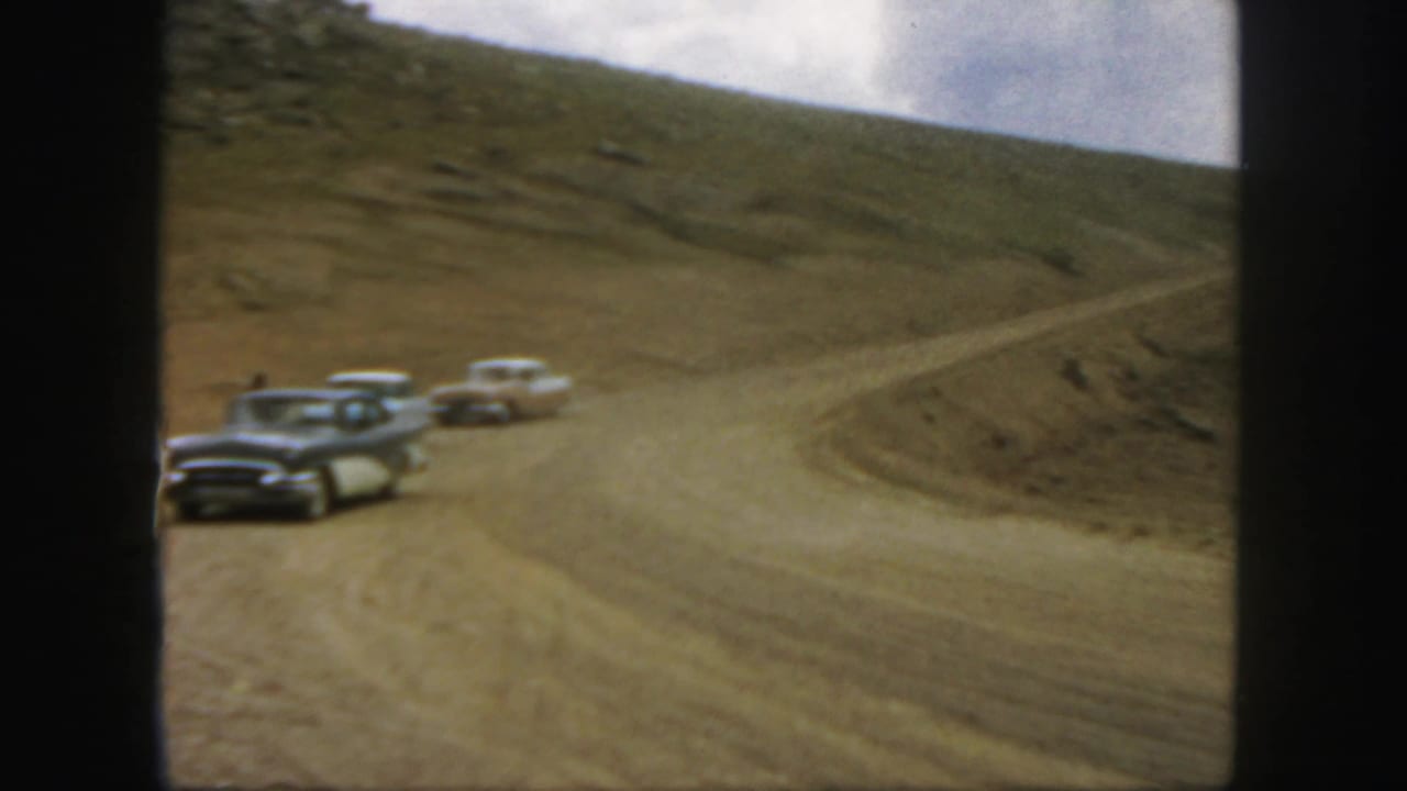 Classic cars drive around a wide dirt curve on a barren mountain road.