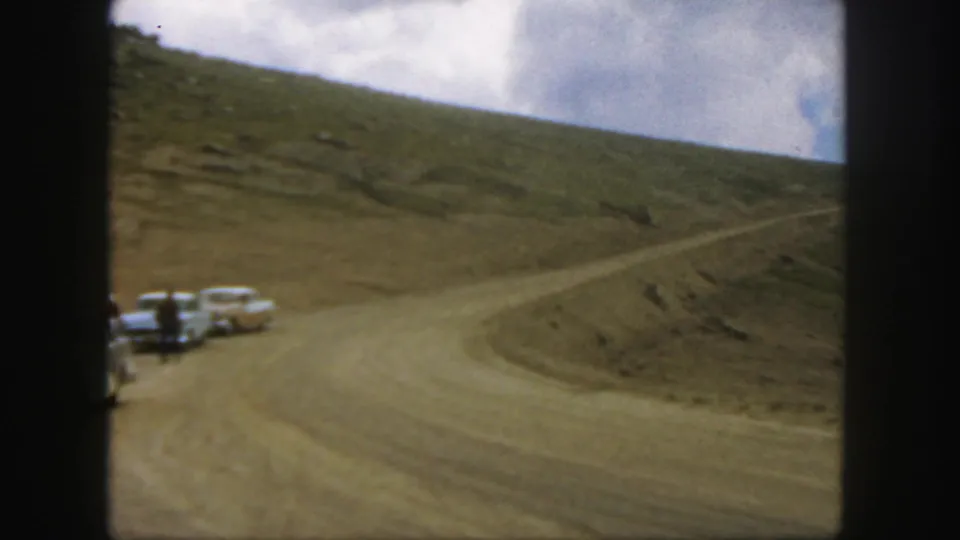 A 1958 Colorado roadside view shows parked classic cars and people beside a winding mountain dirt road.