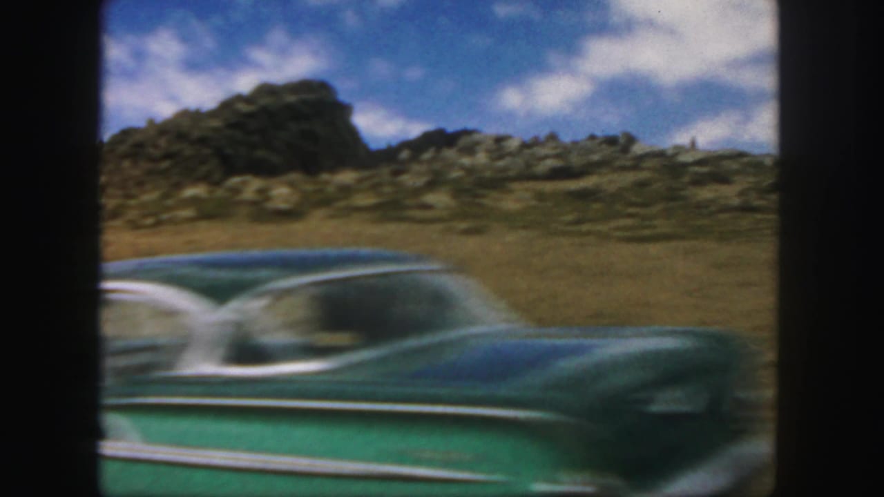 Green classic car passing rocky roadside hills under a blue sky.