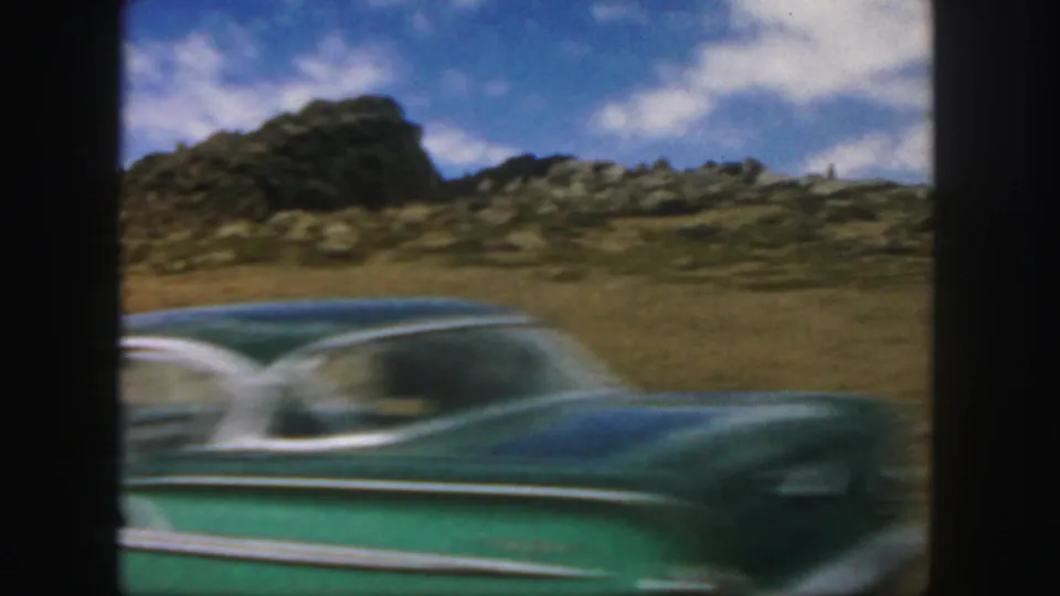 A green classic car passes across a rocky Colorado roadside landscape in 1958 home-movie footage.