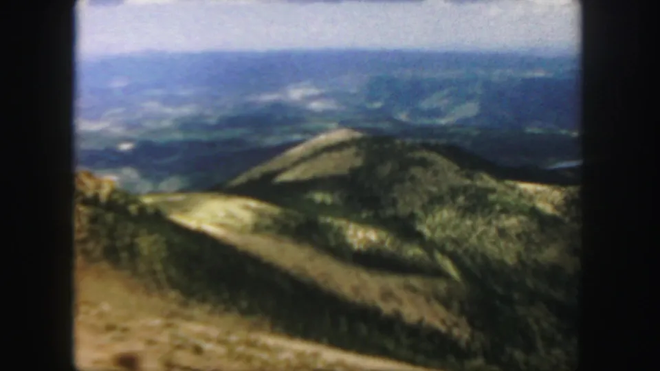 The camera frames a Colorado mountain overlook with wooded slopes, dry foreground, and distant blue ridges.