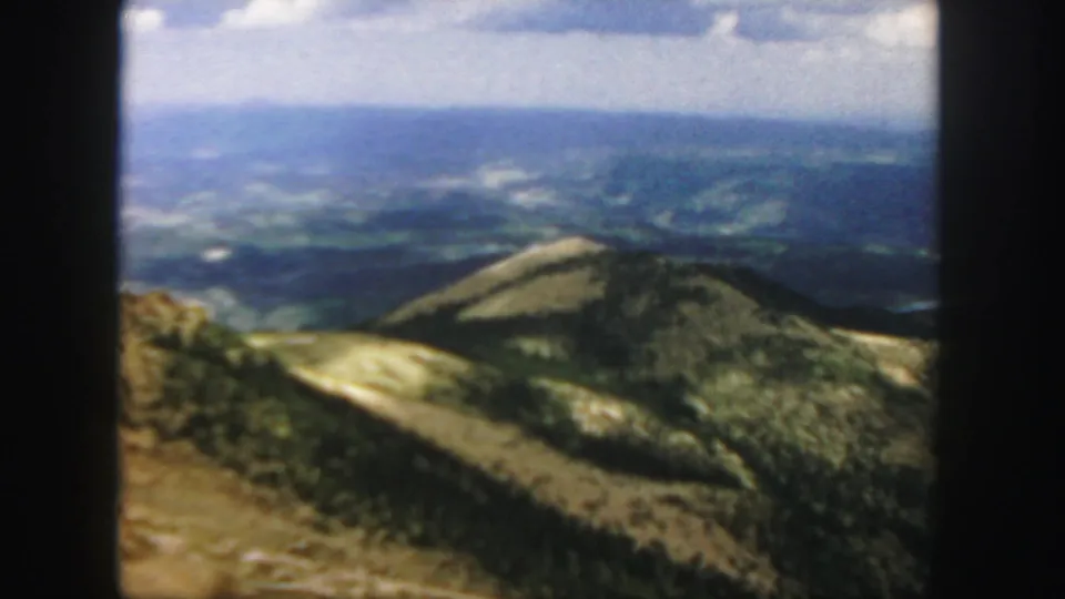 A wide 1958 home-movie view shows a rounded forested hill and layered Colorado mountain ranges beyond.