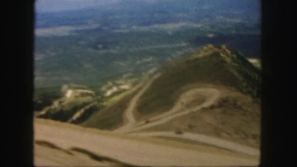 1958 Colorado 8mm frame showing a curved dirt road on an open slope with forest patches and distant blue haze.