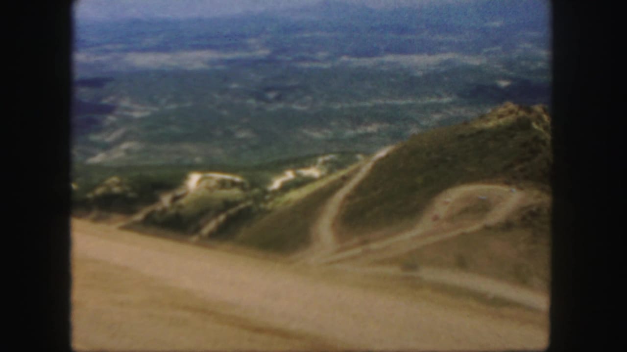 Dusty winding mountain road crossing rounded slopes above a distant valley.