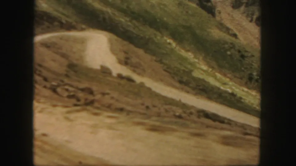 1958 Colorado 8mm frame with a winding dirt road cutting across rocky terrain beneath green mountain slopes.