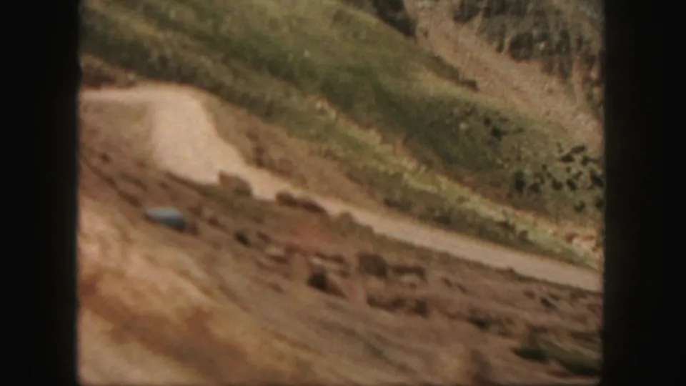 1958 Colorado home-movie frame showing a curving dirt road, rocky roadside, and steep green mountain slope.