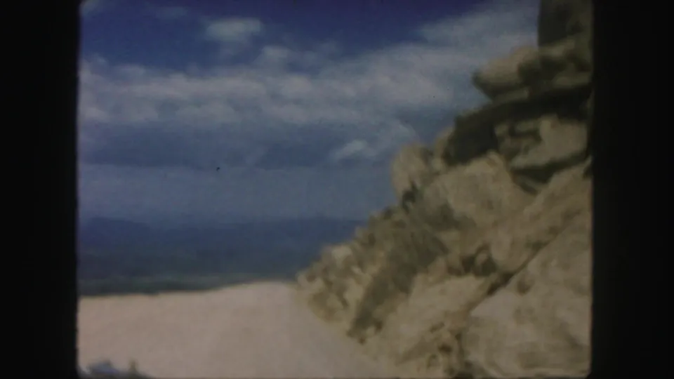 1958 Colorado 8mm frame showing a rocky roadside, pale dirt road, distant hills, and clouds from a moving vehicle perspective.
