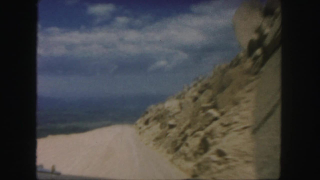 1958 Colorado home-movie frame showing a dirt road passing a rocky mountain slope beneath blue sky and scattered clouds.