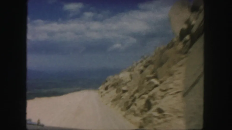 1958 Colorado home-movie frame showing a dirt road passing a rocky mountain slope beneath blue sky and scattered clouds.