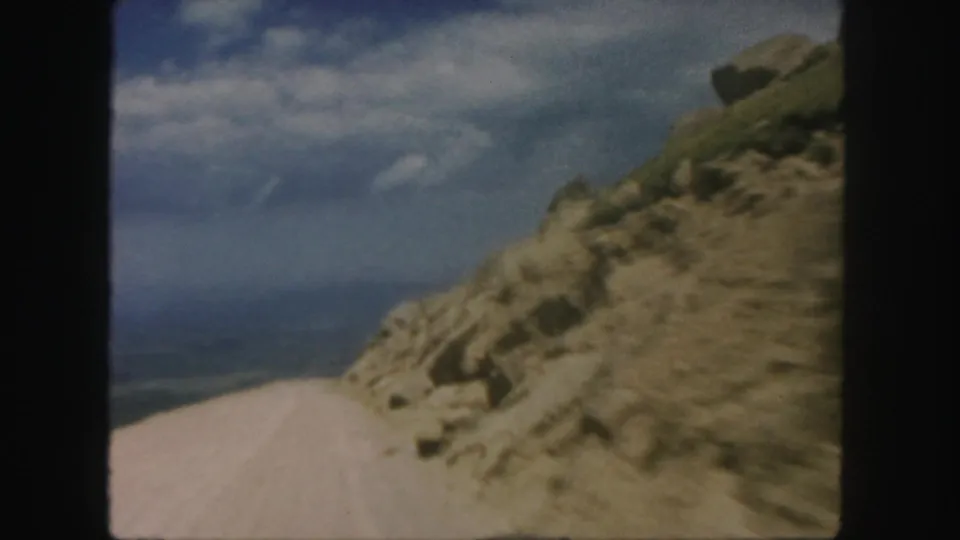 1958 Colorado travel footage frame with an unpaved road running past rough rocks toward distant mountain views.