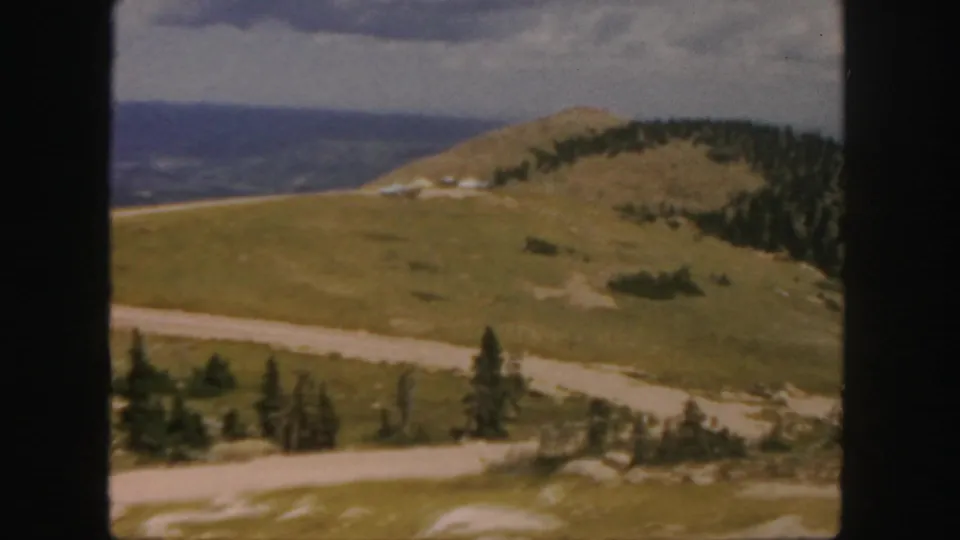 1958 Colorado home-movie frame looking across grassy hills, a winding road, pine trees, and hazy distant ridgelines.