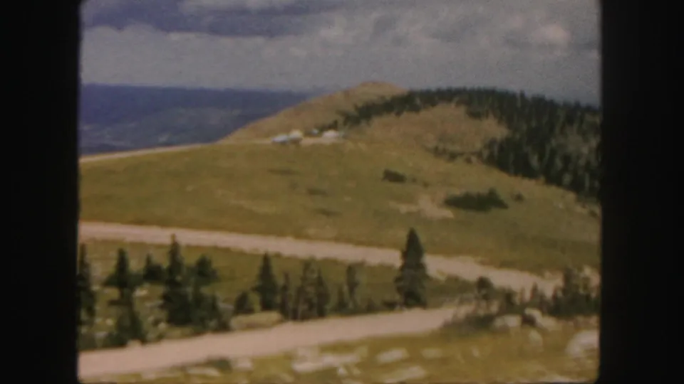 1958 Colorado frame showing a curving dirt road below a grassy hill, with pines and distant mountain ridges in view.