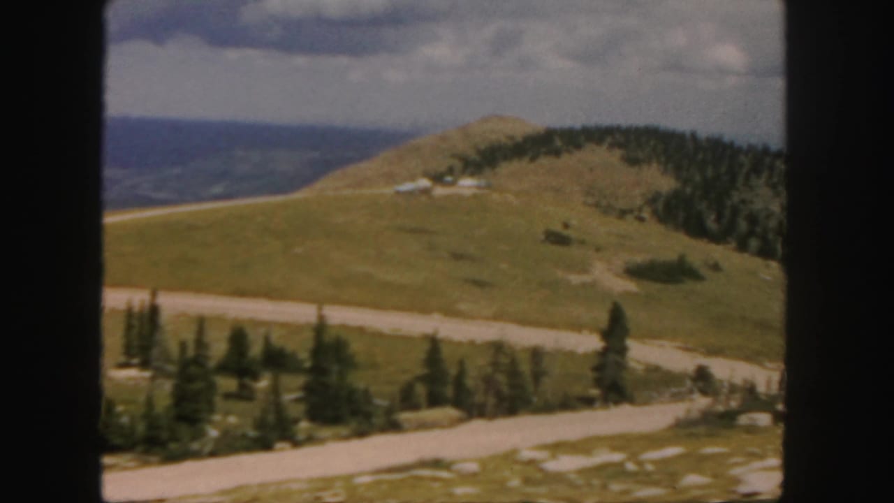 Green mountain hillside with winding road, evergreen trees, distant ridges, and cloudy sky.
