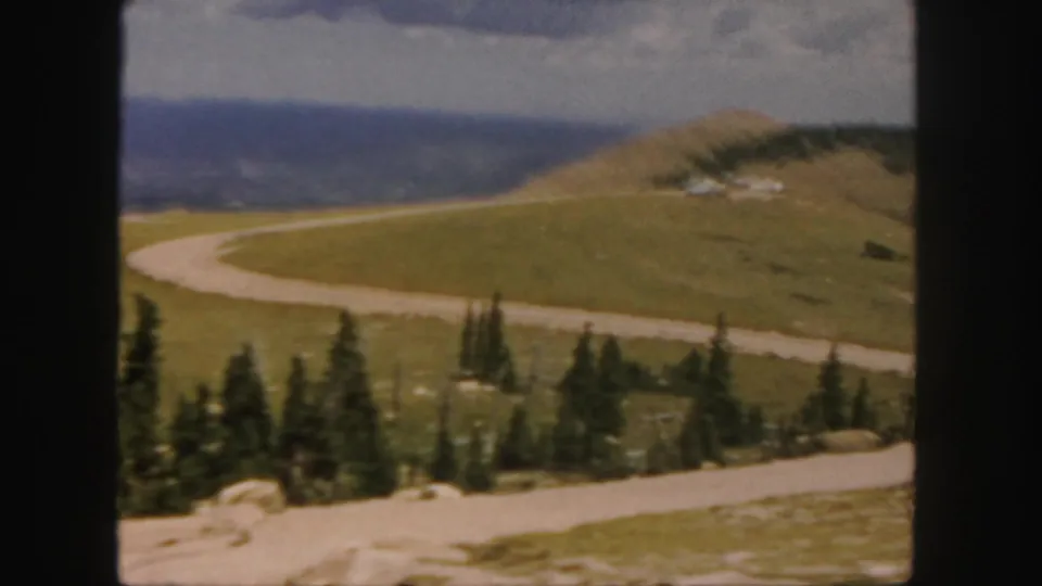 1958 Colorado home-movie view of a pale road curving through grassy mountain terrain with evergreens and blue distant ridges.