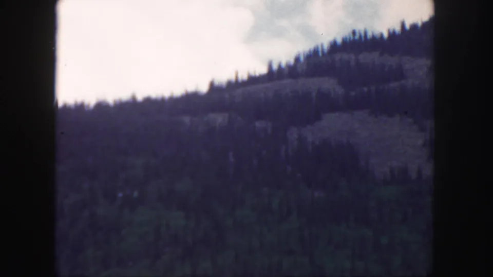1958 Colorado 8mm frame showing a darker forested mountain slope, pale sky, and the edge of the passing landscape view.