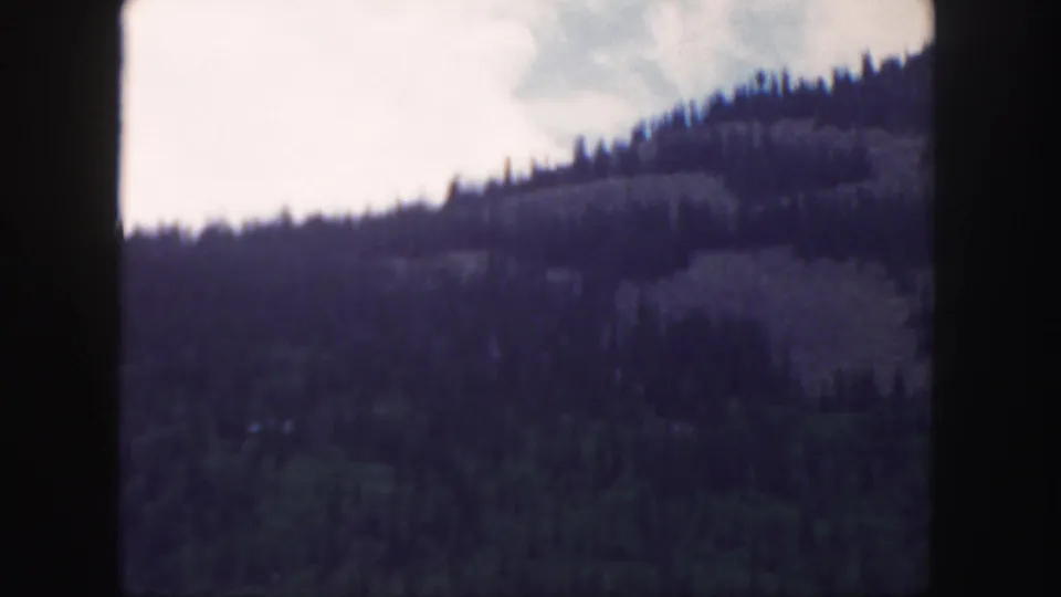 1958 Colorado scenic frame with evergreen-covered hills rising toward a bright sky, softened by grain and faded color.