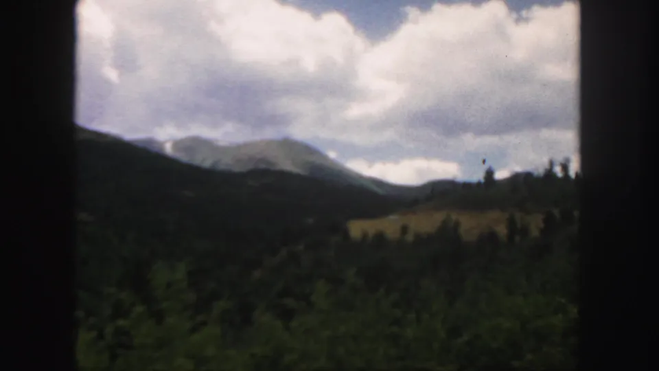 1958 Colorado frame showing a distant mountain peak, rolling forested hills, and clouds above the landscape.