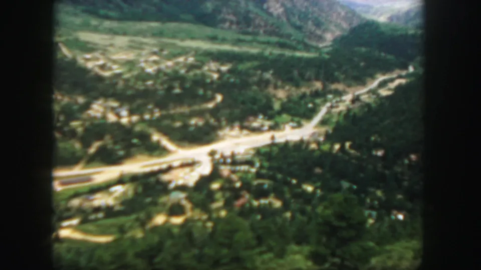 A soft 8mm aerial frame shows a Colorado mountain valley with roads, forest, and a small settlement below.