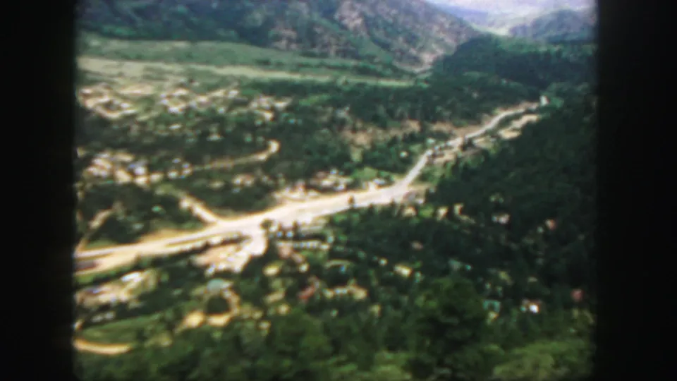 1958 Colorado home-movie frame from above a wooded valley, with winding roads and distant mountain ridges.
