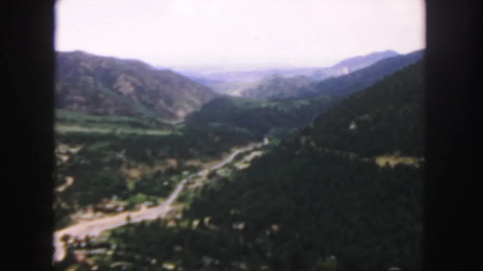 1958 Colorado 8mm view across a wooded mountain valley, with roadways below and hazy ridgelines under a bright sky.