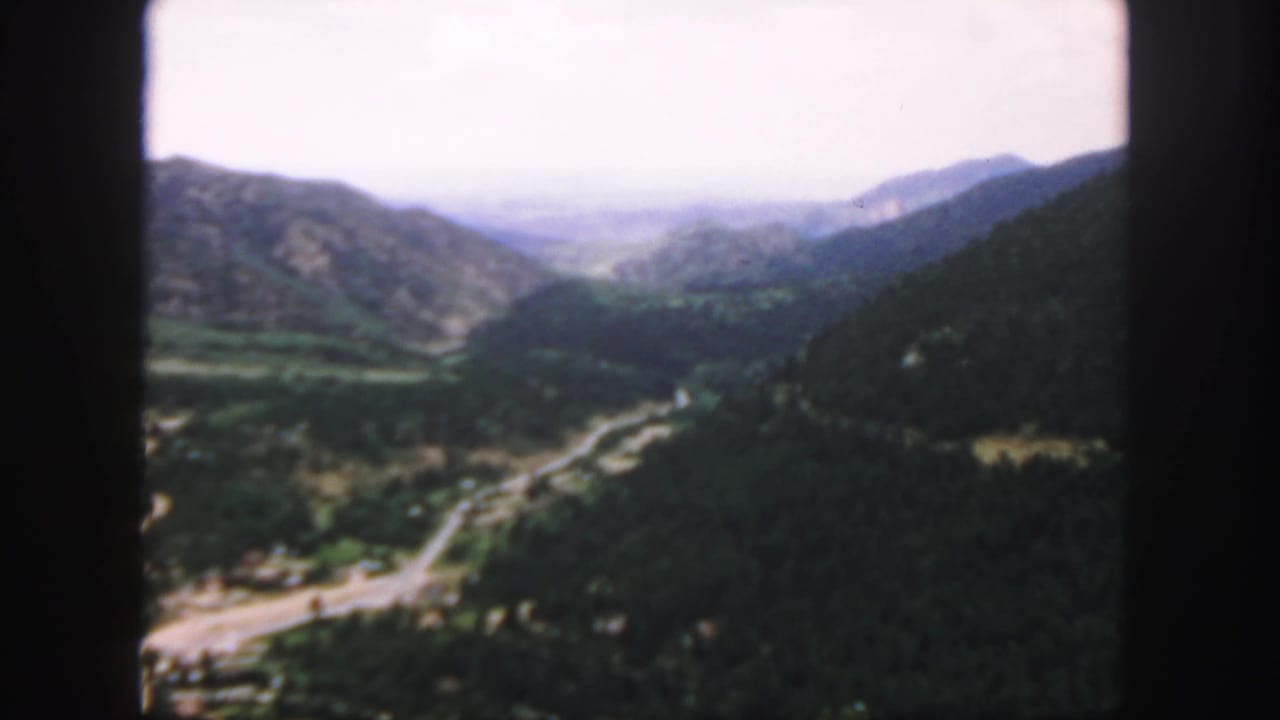 1958 Colorado home-movie frame looking over a mountain valley with a winding road, wooded slopes, and distant ridges.