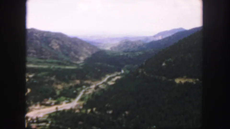 1958 Colorado home-movie frame looking over a mountain valley with a winding road, wooded slopes, and distant ridges.