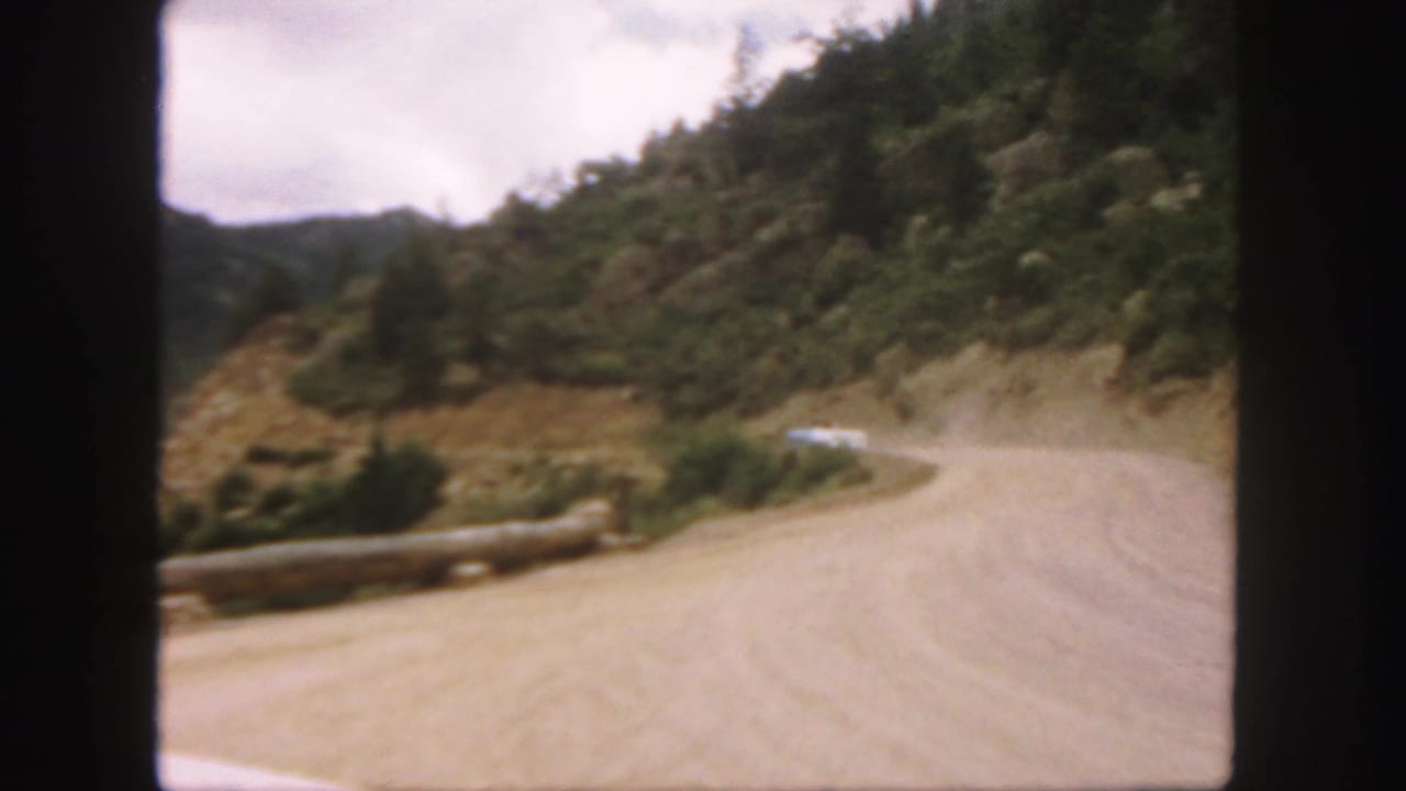 Dusty mountain road curves along a hillside with a pale car ahead and trees on the slope.