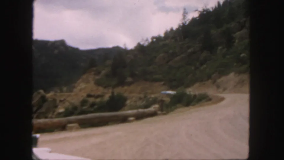 Colorado 1958 home-movie frame of a curving dirt road, timber roadside barrier, hillside trees, and a pale car ahead.