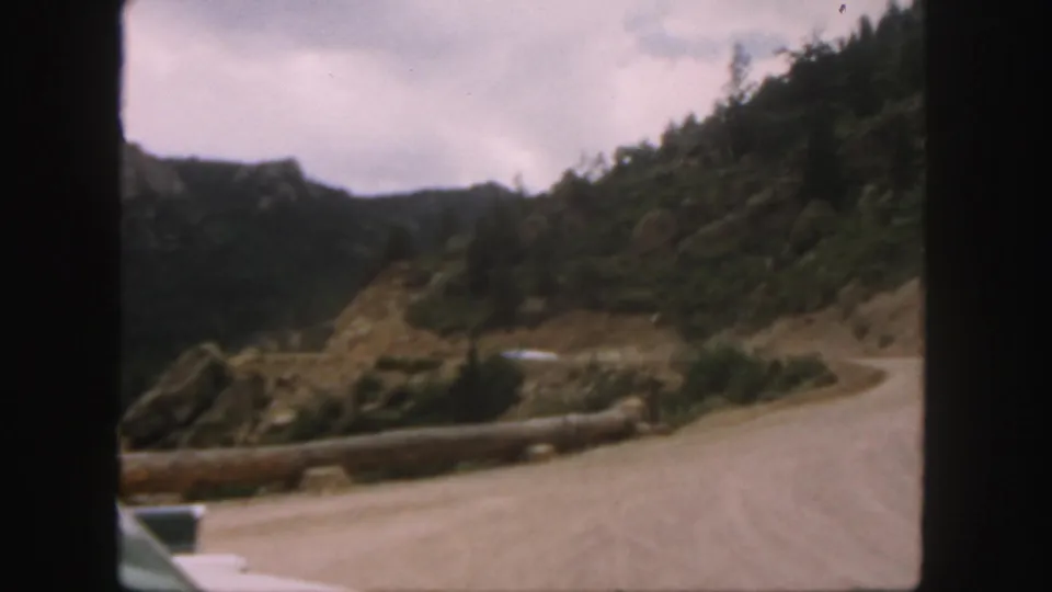1958 Colorado home-movie frame showing a winding dirt road, pale car, log barrier, and forested mountain backdrop.