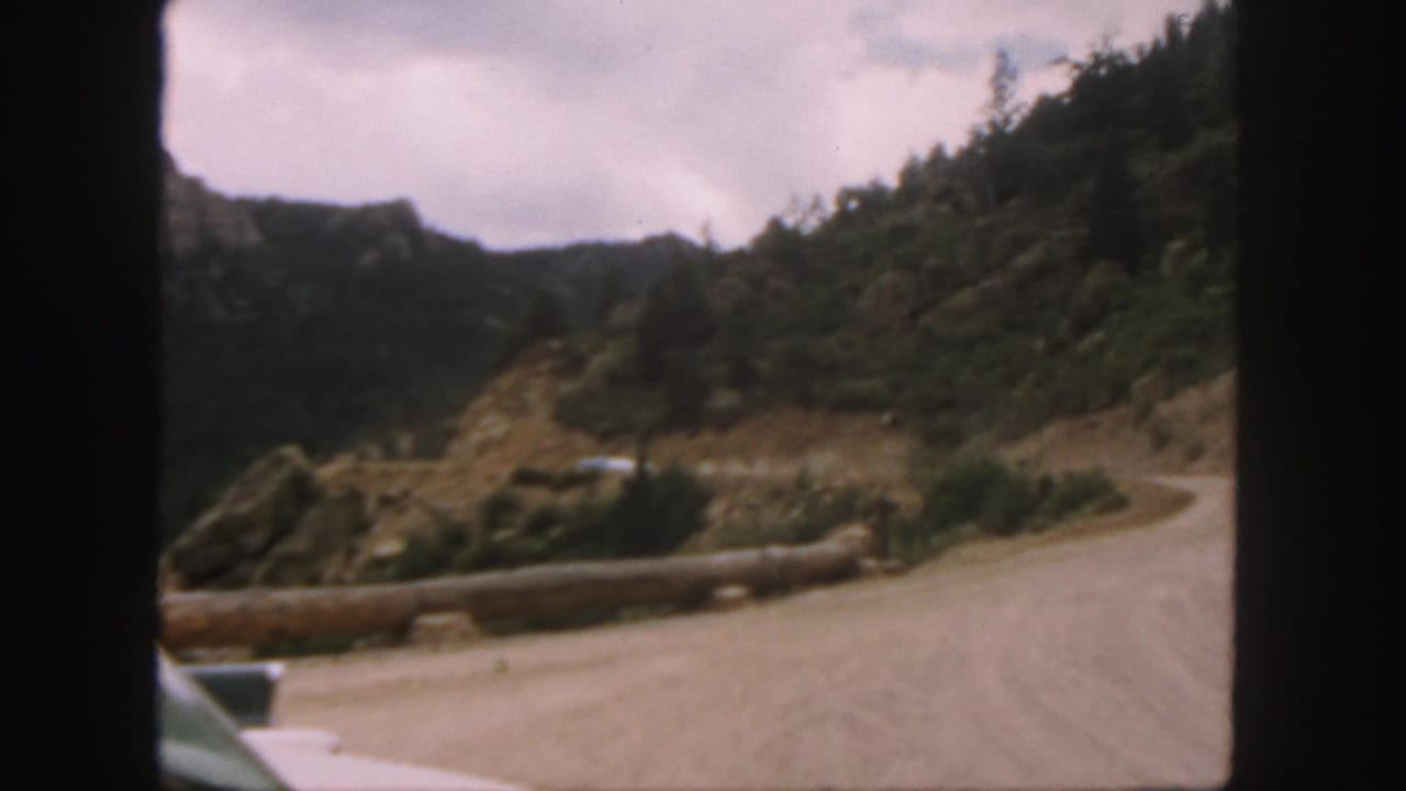 1958 Colorado home-movie frame showing a winding dirt road, log barrier, rocky hillside, and forested mountain slopes.