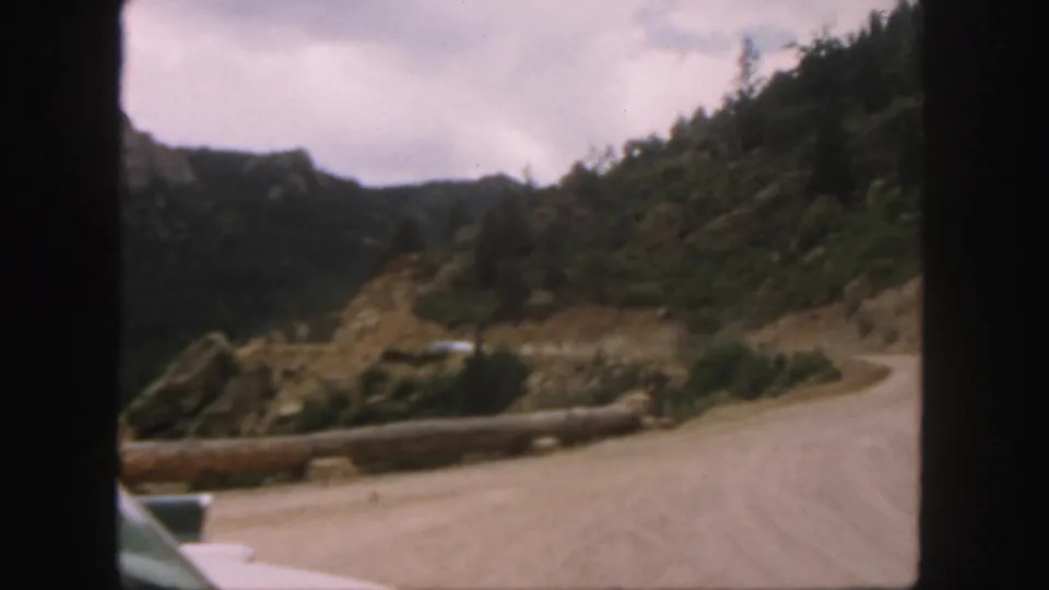 1958 Colorado home-movie frame showing a winding dirt road, log barrier, rocky hillside, and forested mountain slopes.