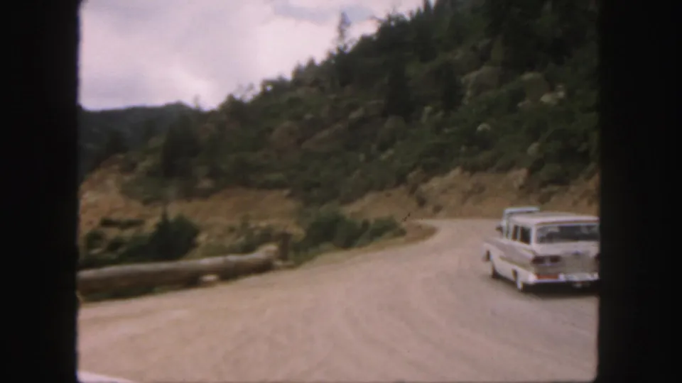 A white classic American car appears on the winding Colorado dirt road, with trees, rocks, and a roadside log barrier nearby.