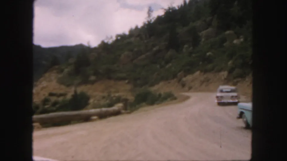 Two mid-century cars are visible near the edge of a winding Colorado dirt road bordered by rocky slopes and evergreen trees.