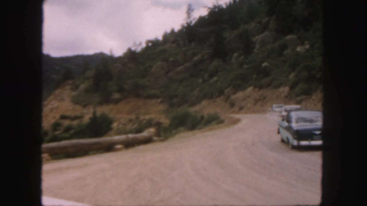 Cars approach on a winding dirt road through a rocky mountain landscape.
