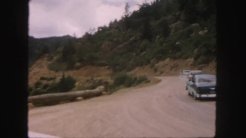1958 Colorado home-movie frame showing cars on a winding unpaved mountain road with evergreen slopes and dark film borders.