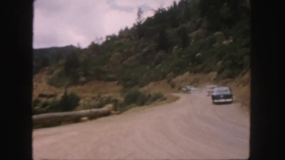 1958 Colorado road-trip frame with a dark sedan on a wide unpaved mountain curve and trees on the hillside.
