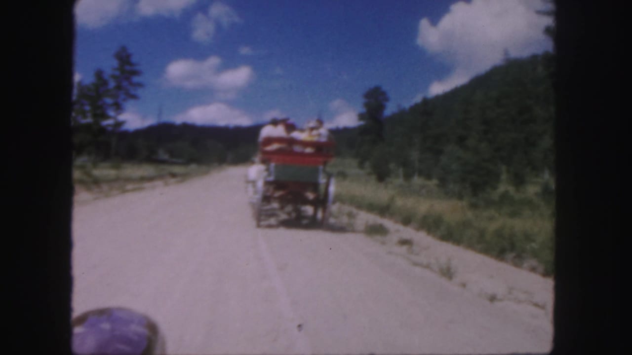 1958 Colorado 8mm frame showing a horse-drawn carriage moving away on a pale dirt road with wooded hills and blue sky.