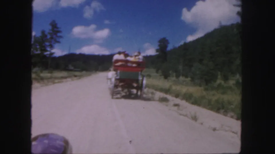 1958 Colorado 8mm frame showing a horse-drawn carriage moving away on a pale dirt road with wooded hills and blue sky.