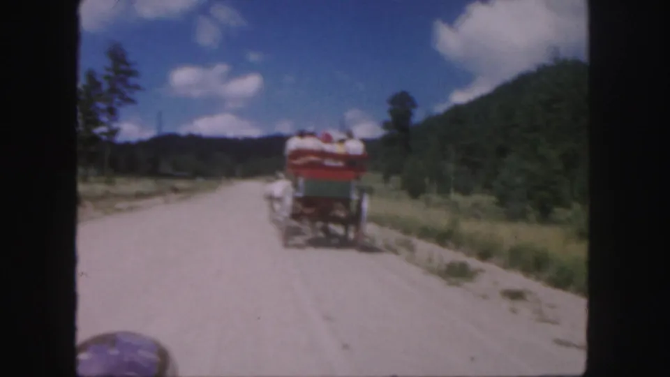 1958 Colorado home-movie frame of a passenger carriage traveling away on a dirt road in a wooded mountain setting.