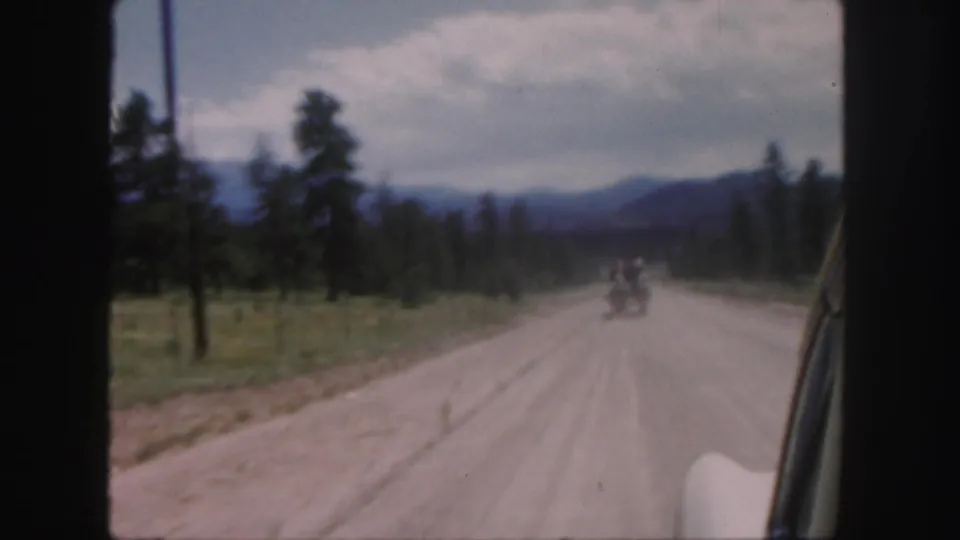 1958 Colorado home-movie frame showing a distant horse-drawn carriage ahead of a car on a rural dirt road.