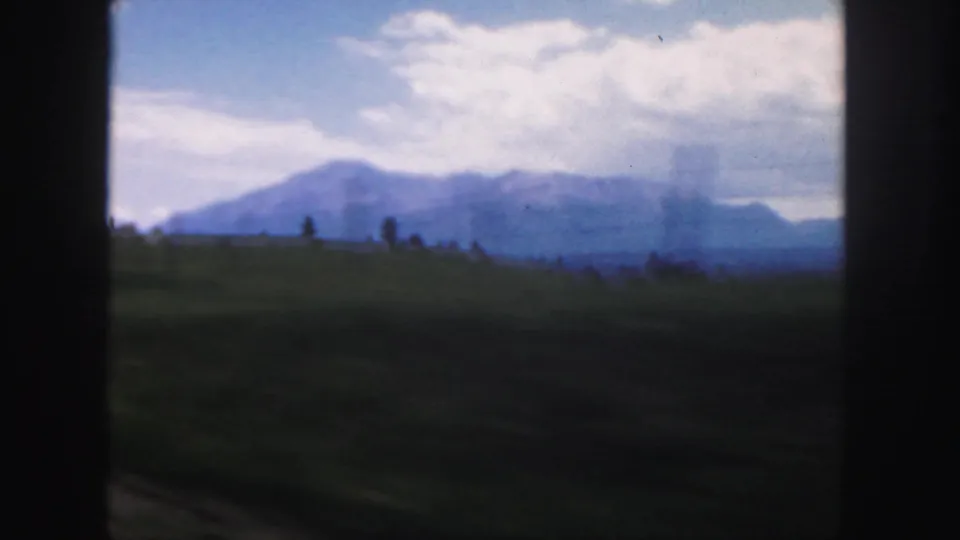 1958 Colorado 8mm frame showing grassy rolling land, small tree shapes, distant blue mountains, and a bright clouded sky.