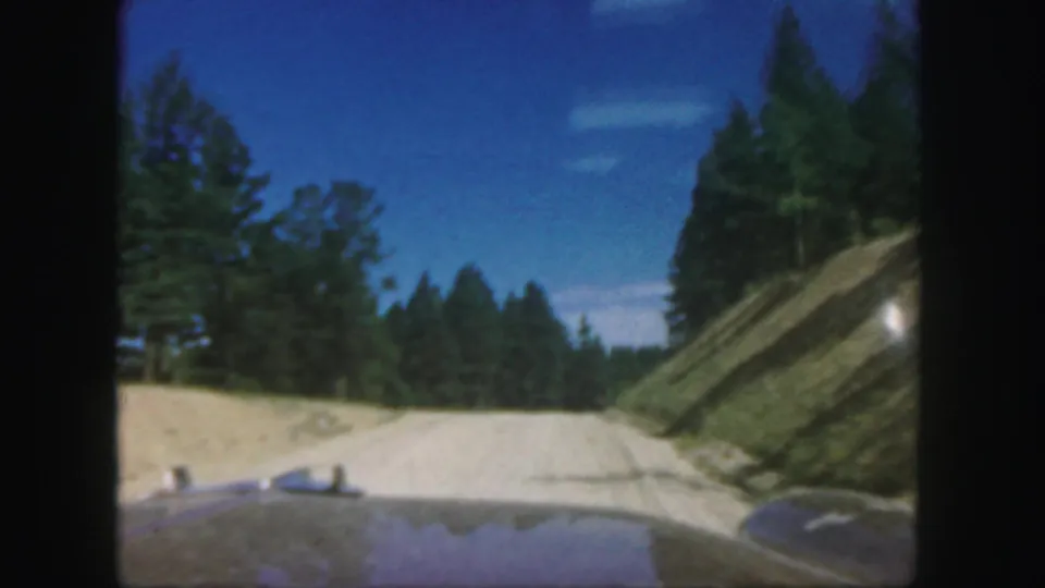 1958 Colorado driving view with car hood, pale dirt road, pine forest, cut hillside, and blue sky in 8mm home-movie color.