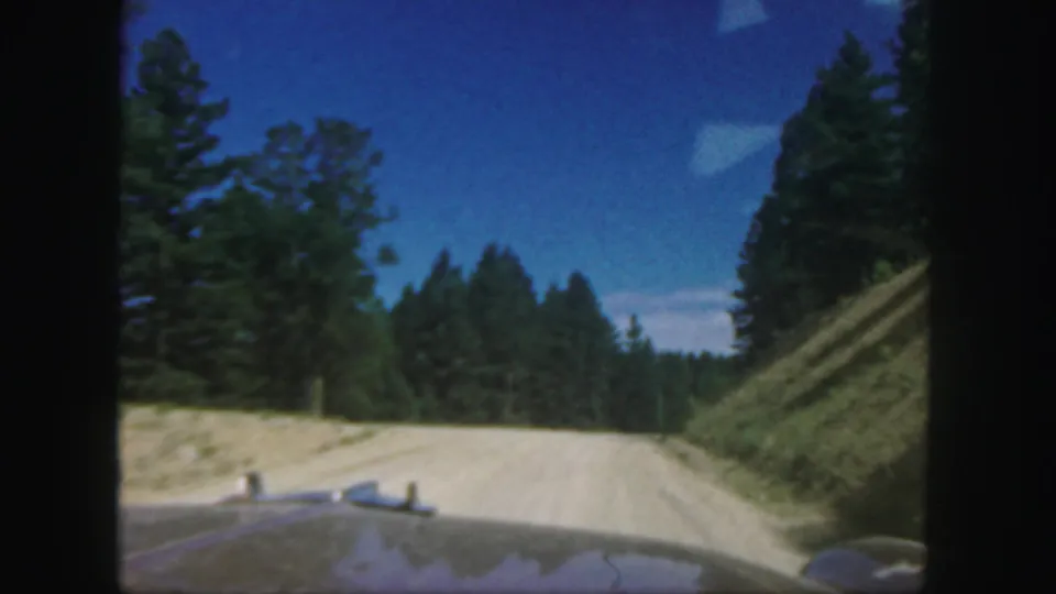 1958 Colorado 8mm frame looking over a vehicle hood along a pale dirt road bordered by pine trees and blue sky.